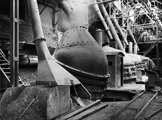 Ein Schwarz-Weiß-Foto einer großen Maschine in einer Fabrik, umgeben von verschiedenen Maschinen, Polen, Treppen, Geländern und anderen Gegenständen, mit Eisenbahnschienen unten.