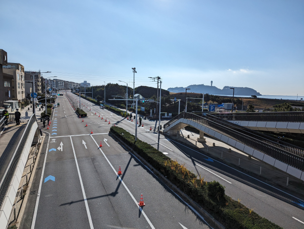 Straße mit Verkehrskegeln, eine Brücke mit Geländern, Laternenpfähle, Gebäude und einige Menschen; Hügel und bewölkter Himmel im Hintergrund, Teil des Takayama-Brücken-Projekts.