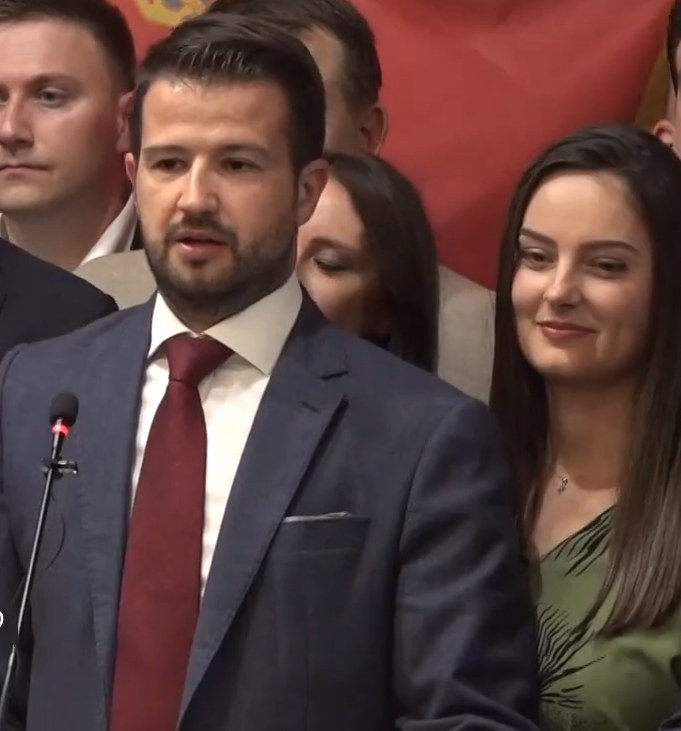 Ein Mann im Anzug spricht vor einem Publikum mit Mikrofon an einem Rednerpult, hinter einem Banner, öffentlich diskutiert über ein Regierungssozialmedienverbot.