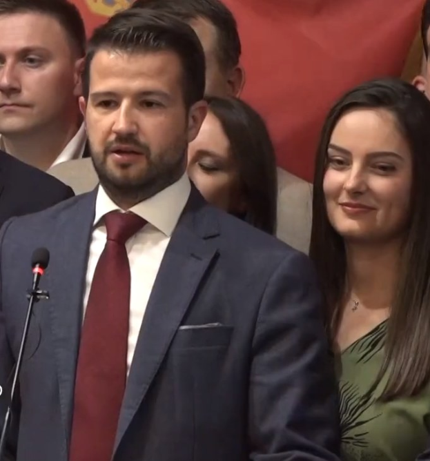 Ein Mann im Anzug spricht vor einem Publikum mit Mikrofon an einem Rednerpult, hinter einem Banner, öffentlich diskutiert über ein Regierungssozialmedienverbot.