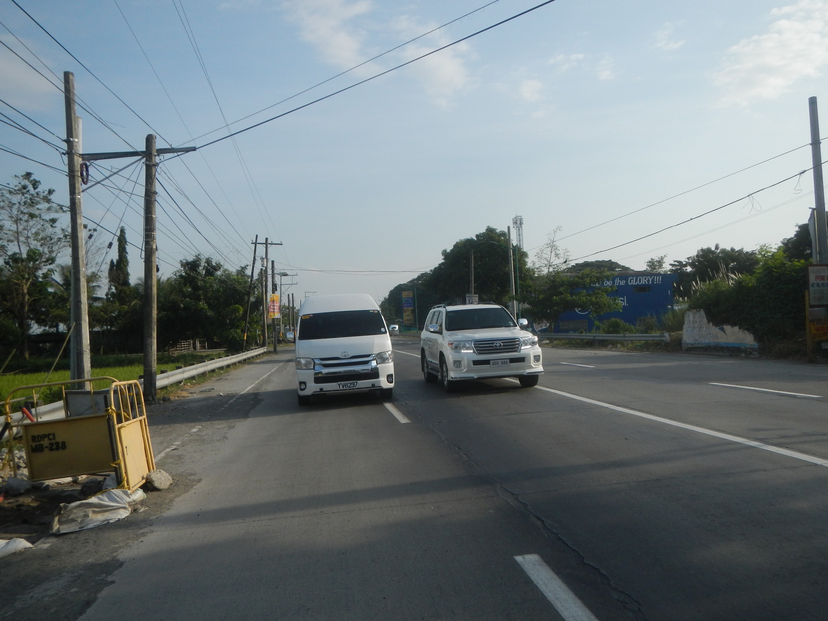 Zwei weiße Lieferwagen fahren auf einer Straße mit Strommasten, Kabeln, einer gelben Absperrung, Bäumen, Gebäuden und einem klaren blauen Himmel im Hintergrund.