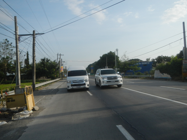 Zwei weiße Lieferwagen fahren auf einer Straße mit Strommasten, Kabeln, einer gelben Absperrung, Bäumen, Gebäuden und einem klaren blauen Himmel im Hintergrund.
