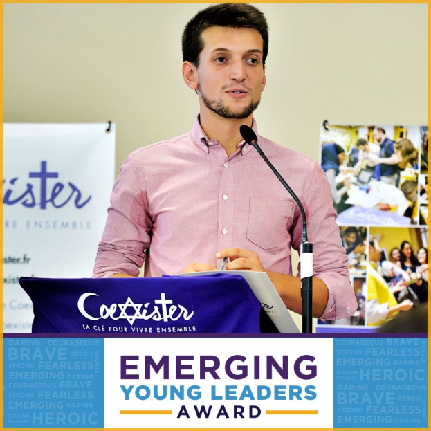 Ein Mann steht an einem Rednerpult mit einem Mikrofon, haltend ein Papier, vor einem Banner mit der Aufschrift "Emerging Young Leaders Award", mit mehreren Personen rechts von ihm.