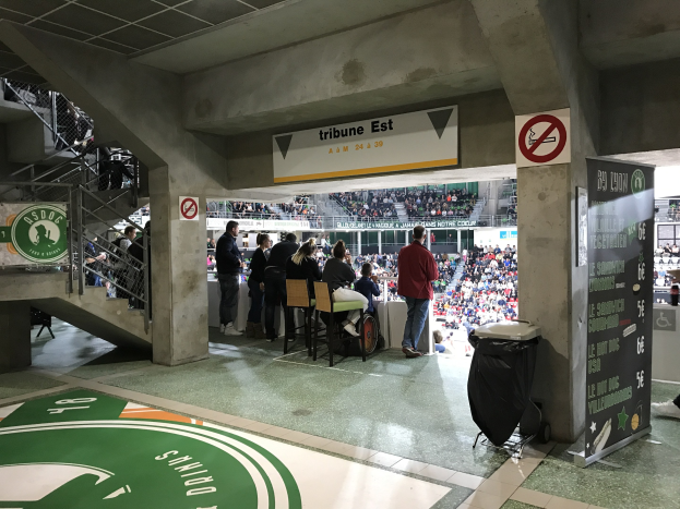 Eine Gruppe von Menschen steht vor einer sitzenden Menge in einem Stadion, mit Schildern, die "Boston Celtics vs New York Jets" anzeigen, einem Müllcontainer, einer Texttafel, einer Treppe mit Geländern und Deckenleuchten.