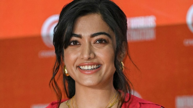 Eine Frau in einem roten Kleid mit einem eleganten Dutt und dezentem Make-up steht lächelnd da, trägt schwarze High Heels und hat ein hellgelbes Banner mit der Aufschrift "Sonam Kapoors schönste Looks vom Cannes Film Festival" im Hintergrund.