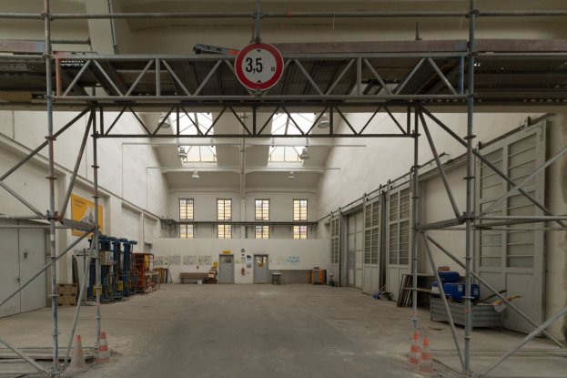 Lagerhalle mit Gerüst, Verkehrskegeln, Metallstangen, einem Schild, Maschinen, einer Tür, Fenstern, Wänden, Deckenleuchten und einem Dach.