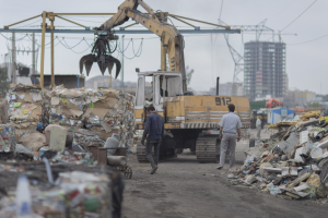 Eine Gruppe von Männern geht auf einem Schotterweg neben einem Müllhaufen, im Hintergrund ist ein Bagger zu sehen, Gebäude, Bäume und andere Objekte sind verteilt.