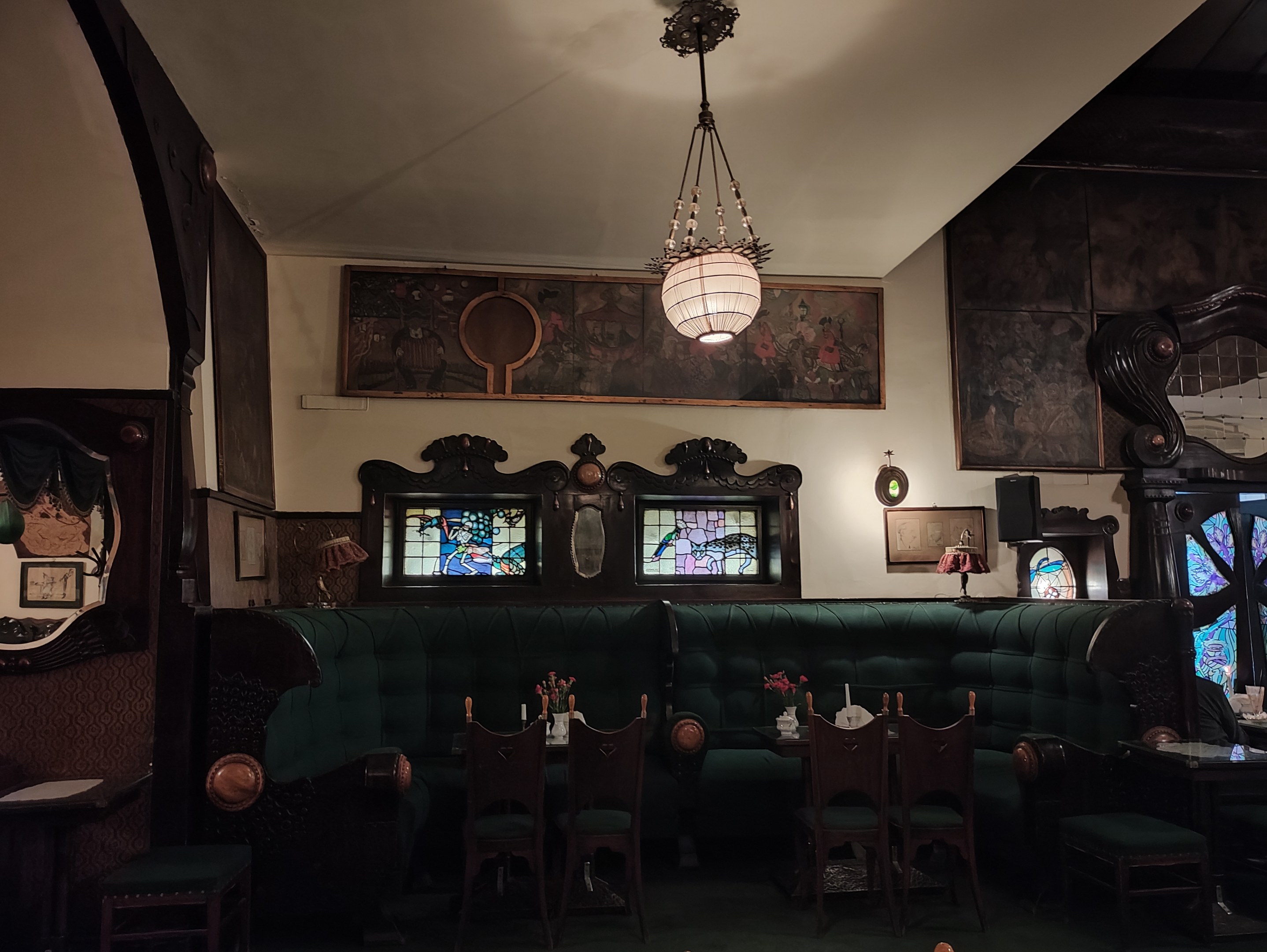 Innenraum eines liebevoll eingerichteten Restaurants mit Tischen, Stühlen, Blumenvasen, Sofas, einem Kronleuchter, gerahmten Kunstwerken an den Wänden und einem Fenster auf der rechten Seite.