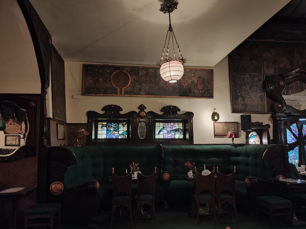 Innenraum eines liebevoll eingerichteten Restaurants mit Tischen, Stühlen, Blumenvasen, Sofas, einem Kronleuchter, gerahmten Kunstwerken an den Wänden und einem Fenster auf der rechten Seite.