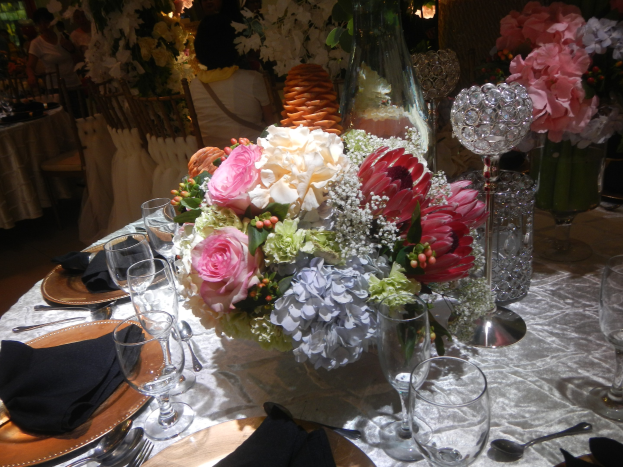 Ein formeller Esstisch mit Tellern, Besteck, Gläsern, Servietten und Blumenvasen, mit einem Blumenarrangement in der Mitte, im Hintergrund Stühle und Tische und einige sitzende Personen.