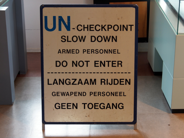 Bodenaufschrift "UN-Checkpoint Slow Down Armed Personnel - Do Not Enter" mit einer Glasplatte und Gegenständen rechts sowie einer Wand mit einem Gegenstand im Hintergrund.