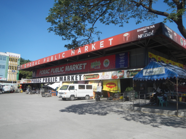 Öffentlicher Markt mit einem weißen Lieferwagen, der davor geparkt ist, umgeben von Gebäuden, Motorfahrzeugen, Menschen, die unter einem Zelt sitzen, Geländern, Bäumen und einem klaren blauen Himmel.