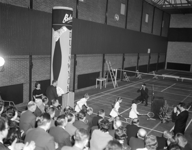 Schwarzes und weißes Foto von Menschen, die Tennis in einer Turnhalle spielen, einige stehen und einige sitzen mit Schlägern, eine Uhr an der Wand im Hintergrund.
