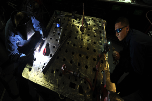 Zwei Männer in Schutzausrüstung arbeiten an einem Metallstück in einer schwach beleuchteten Werkstatt, umgeben von verschiedenen Werkzeugen und Materialien auf einem Tisch.