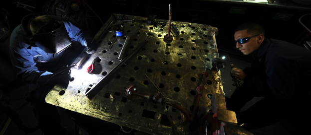 Zwei Männer in Schutzausrüstung arbeiten an einem Metallstück in einer schwach beleuchteten Werkstatt, umgeben von verschiedenen Werkzeugen und Materialien auf einem Tisch.