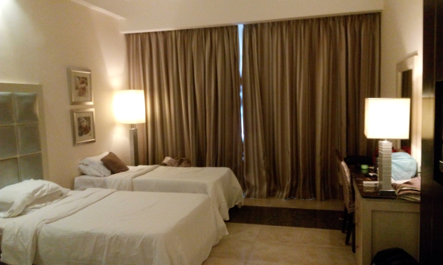 Hotelzimmer mit zwei Betten, jeweils mit weißen Laken und Kissen, Holzschrank, Teppichboden, Stuhl, Tisch mit Gegenständen, Spiegel, Fotos auf der Wand und brauner Gardine.