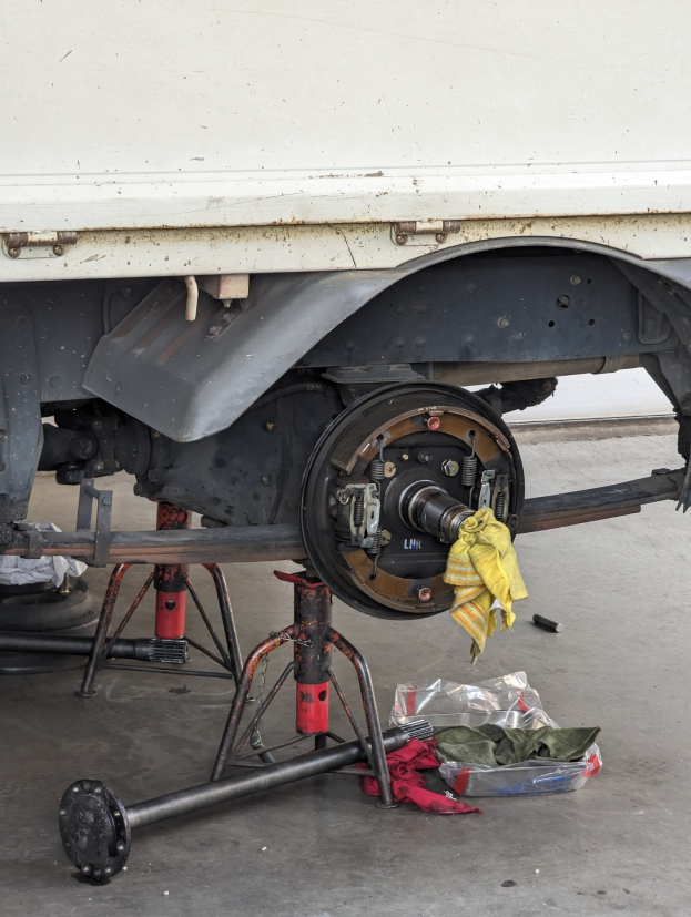Ein gelbes Tuch liegt über einem in einer Garage geparkten Lkw, mit Werkzeugen und Gegenständen, die vor ihm auf dem Boden verstreut sind.