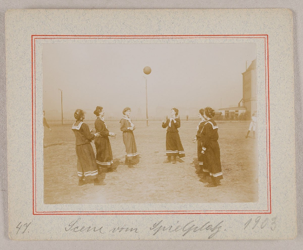 Eine Gruppe von Frauen, die auf dem Boden Fußball spielen, mit einem Ball in der Mitte, Pfosten und Gebäuden im Hintergrund und einem klaren blauen Himmel, mit Text am unteren Rand des Bildes.