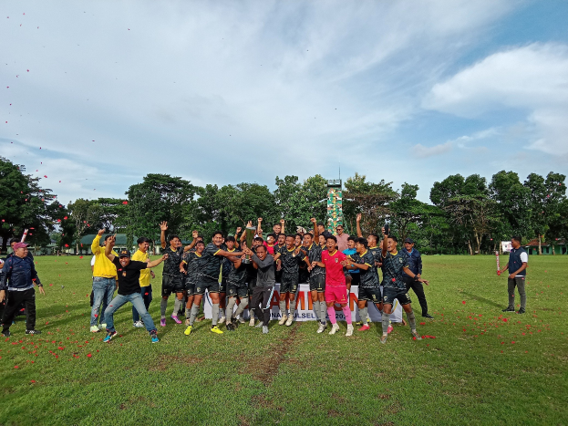 Eine Gruppe von Menschen auf einem grünen Feld umgeben von Bäumen und einem klaren blauen Himmel mit einem Banner im Hintergrund, auf dem "Cambodia Rugby World Cup 2019" steht.