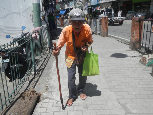 Ein älterer Mann geht die Straße entlang, hält in einer Hand einen Stock, in der anderen eine Tasche und einen Stock und trägt eine Mütze, mit einem Metallzaun links und Fahrzeugen, Gebäuden und Schildern im Hintergrund.
