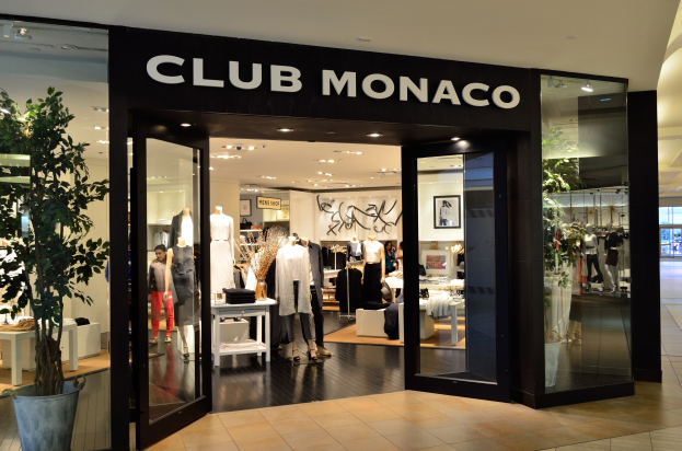 Vorführfläche eines Club Monaco-Geschäfts in einem Einkaufszentrum mit Glaswänden und -türen, Schaufensterpuppen in Kleidern, Auslagetischen mit Waren, einem Topf mit einer Pflanze und Deckenbeleuchtung.