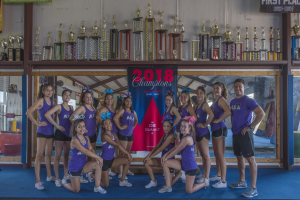 Eine Gruppe von Cheerleadern in lila T-Shirts und schwarzen Shorts posiert vor einem Vitrinenschrank und einem Banner mit der Aufschrift "2018 Summit Cheerleading Champions."