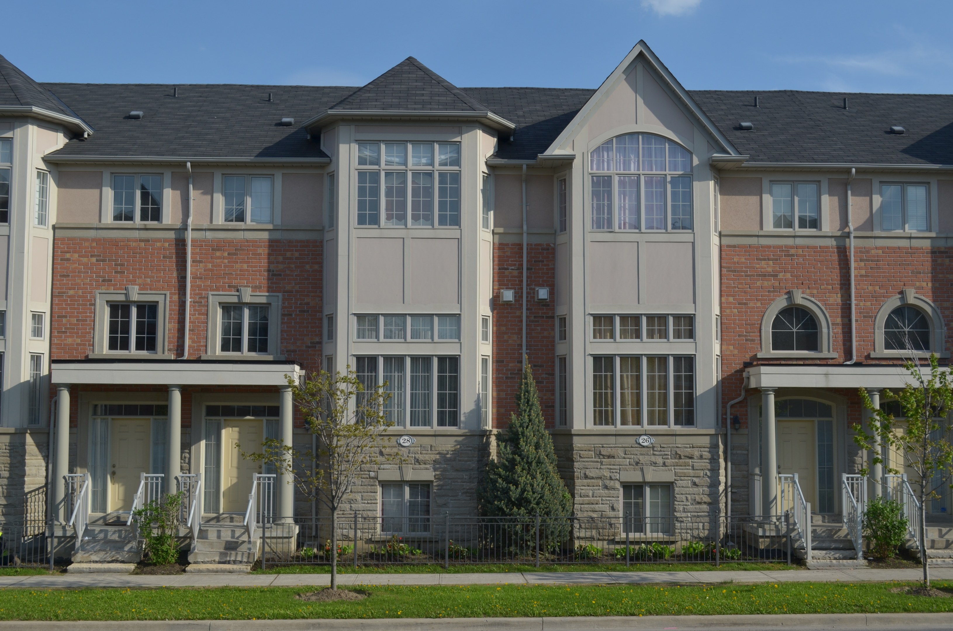 Eine Reihe von Stadthäusern in einer Wohngegend mit Fenstern, Türen, Säulen, Geländern, Stufen, Pflanzen, Bäumen, Gras, einer Straße im Vordergrund und dem Himmel im Hintergrund.
