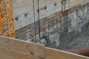 Baustelle mit einem Kran auf der linken Seite, einer betonbedeckten Wand im Hintergrund, einem hölzernen Objekt und einem Rohr unten.