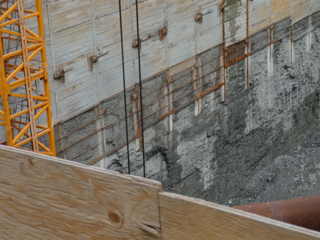Baustelle mit einem Kran auf der linken Seite, einer betonbedeckten Wand im Hintergrund, einem hölzernen Objekt und einem Rohr unten.