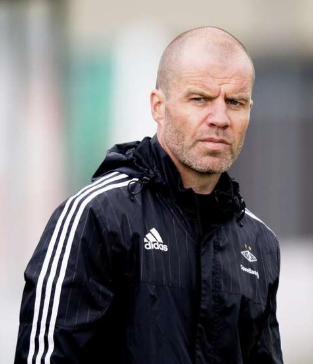 Ein kahlköpfiger Mann in einer schwarzen Jacke steht auf einem Fußballfeld, mit den Händen verschränkt und einem ernsten Gesichtsausdruck.