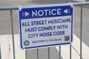 Anschlagtafel an einem Zaun mit einem Schild, auf dem steht "Anschlag: Alle Straßenmusiker müssen die Stadtlärmvorschrift einhalten" und eine Straße im Hintergrund.