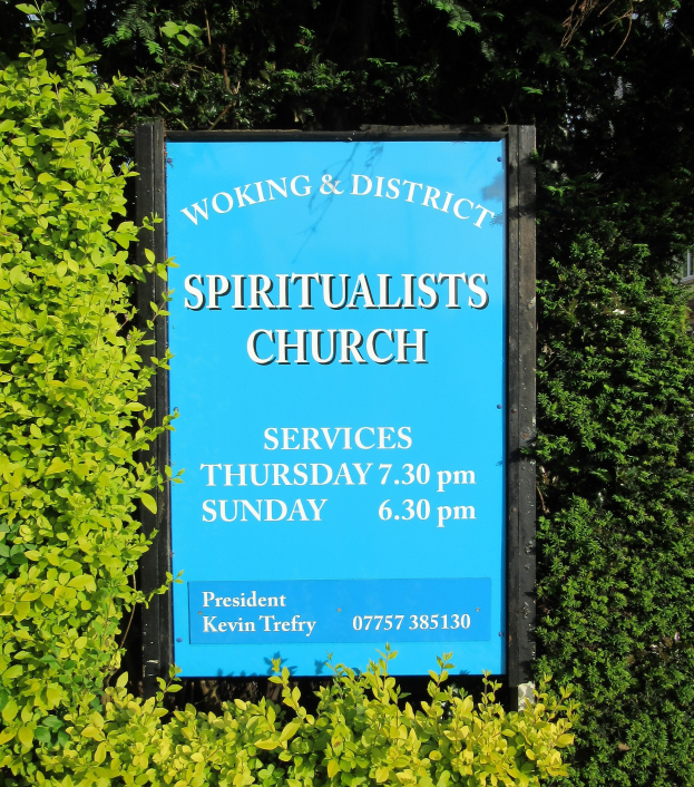 Ein Holzbrett mit der Aufschrift "Woking & District Spiritualists Church" steht draußen, umgeben von Grün mit einem Gebäude im Hintergrund.