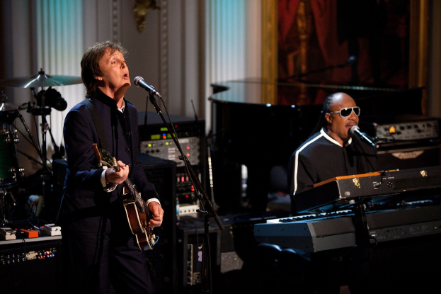 Paul McCartney und Stevie Wonder auf der Bühne performend, McCartney spielt Gitarre und singt, Wonder trägt eine Brille und spielt Piano.