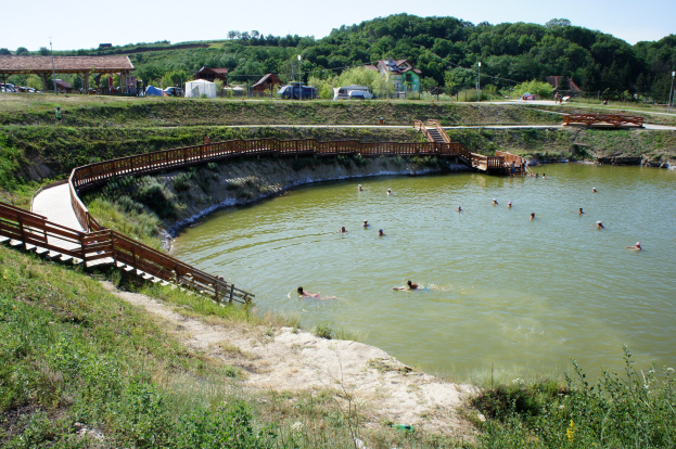 Gruppe von Menschen, die in einem Gewässer schwimmen, umgeben von Grünflächen, einer Brücke mit Treppen zum Wasser und Hütten, Fahrzeugen und Pfählen im Hintergrund unter einem klaren blauen Himmel.