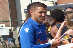 Ein Mann in einem blauen Hemd signiert Autogramme für eine Gruppe von Menschen, einige tragen Mützen und Schutzbrillen, mit einem Gebäude, einem Maschendrahtzaun und einem Fahrrad im Hintergrund.