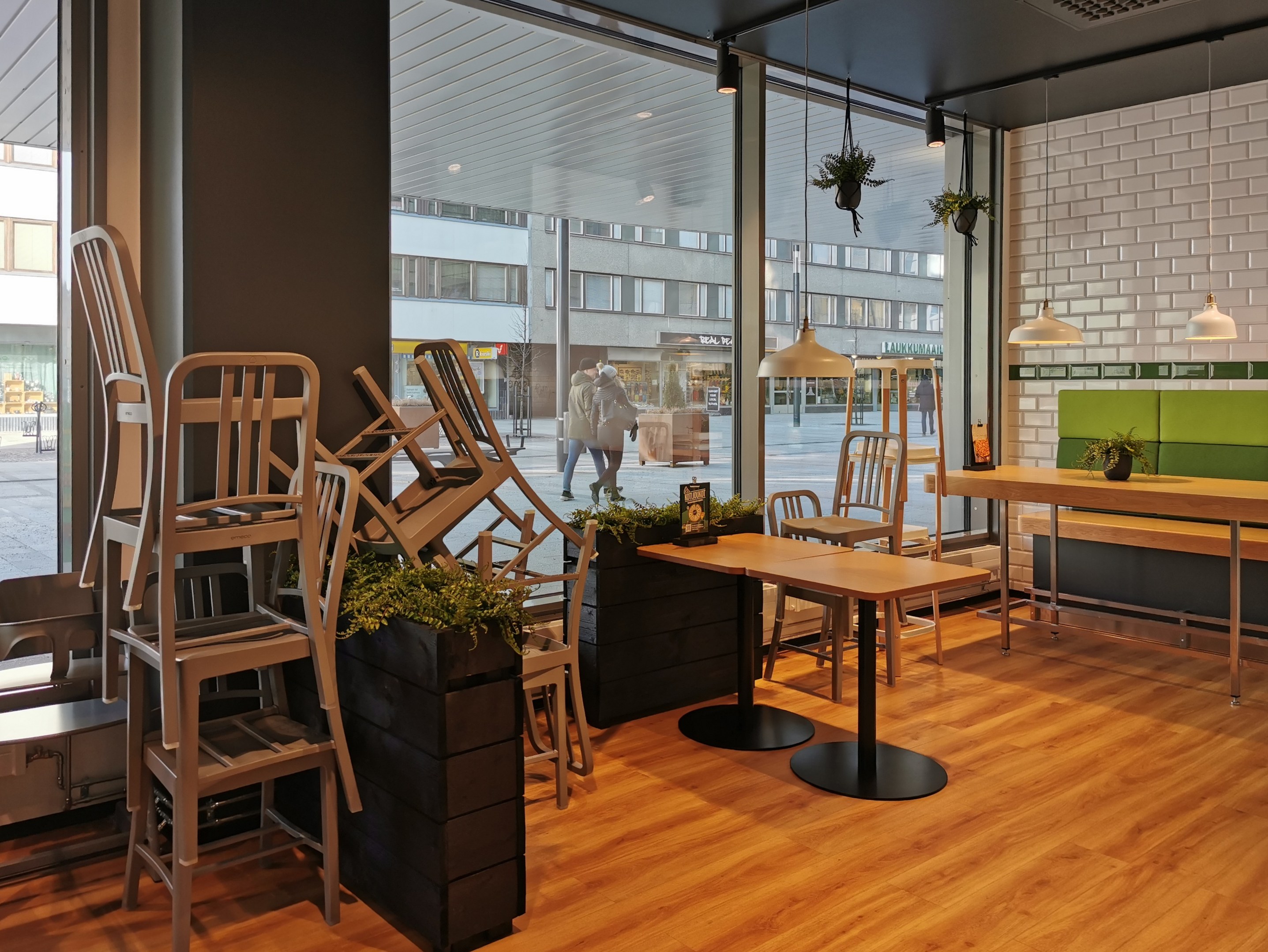 Ein Berliner Restaurant mit hölzernen Tischen und Stühlen vor einem großen Fenster, das eine Außenansicht von Fußgängern, Gebäuden und städtischen Elementen zeigt.