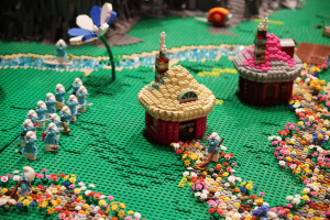 Ein Lego-Modell einer kleinen Stadt mit Blumen, Spielzeugen, Häusern, Pflanzen und anderen Gegenständen vor einem leicht unscharfen Hintergrund.