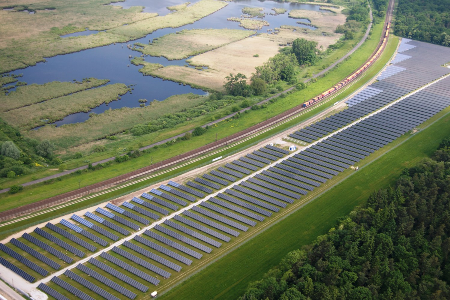 Luftaufnahme einer Solaränlage mit Solarpanelen in einem Feld, umgeben von Bäumen, Gras, Pflanzen und Wasser, mit einem Zug auf einer näheren Eisenbahnschiene.