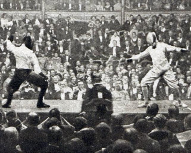 Zwei Männer fechten in einem Stadion, einer hält ein Schwert und der andere einen Schild, mit einer jubelnden Menge und Pfosten im Hintergrund.