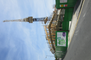 Eine Baustelle mit einem grünen Gebäude im Bau, einer Straße auf der rechten Seite und einem Turm im Hintergrund unter einem bewölkten Himmel.