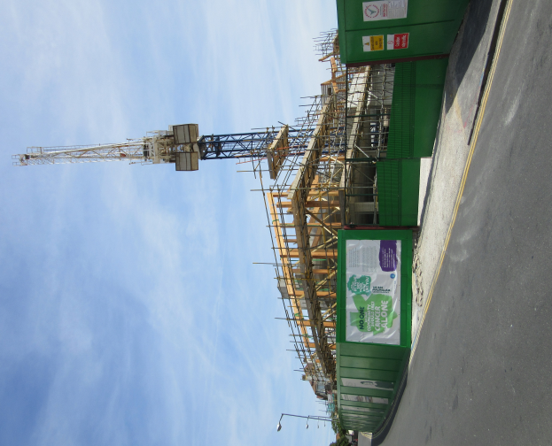 Eine Baustelle mit einem grünen Gebäude im Bau, einer Straße auf der rechten Seite und einem Turm im Hintergrund unter einem bewölkten Himmel.