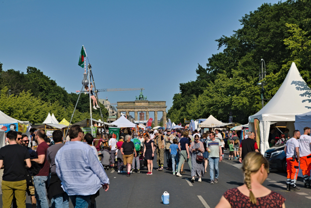 Eine Menge geht eine Straße entlang, gesäumt von Zelten, Fahrzeugen und Bäumen, auf einen Bogen unter einem klaren blauen Himmel zu, mit Fahnenmästen auf der linken Seite, was auf das Oktoberfest in München, Deutschland, hindeutet.