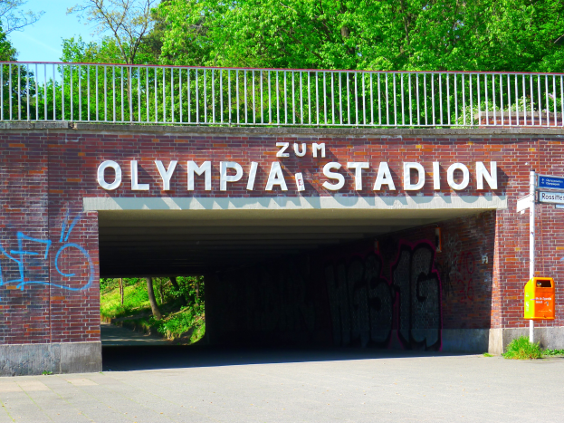 Der Eingang zum Olympiastadion in Berlin, Deutschland, mit einer Brücke mit Text, einem Metallzaun, einer Tafel, einer Kiste, Pflanzen, Gras, einer Baumgruppe und einem bewölktem Himmel.