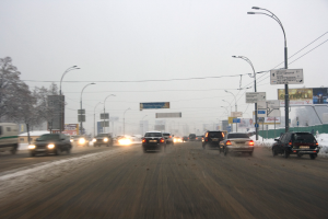 Eine verkehrsreiche Stadtstraße an einem verschneiten Tag mit Fahrzeugen, schneebedeckter Straße, Laternen, Texttafeln, Bäumen, Gebäuden und einem Himmel im Hintergrund.