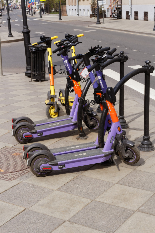 Eine Reihe von Elektrokleinstfahrzeugen auf einem Bürgersteig geparkt mit einem Mülleimer im Vordergrund, vor Gebäuden, Bäumen und Fahrzeugen auf einer Straße.