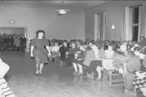 Eine Frau in einem schwarzen und weißen Kleid läuft über einen Laufsteg während einer Modenschau für Frauenrechte, mit einer Menge, die auf Stühlen sitzt und einer Uhr und Lampen an den umliegenden Wänden.