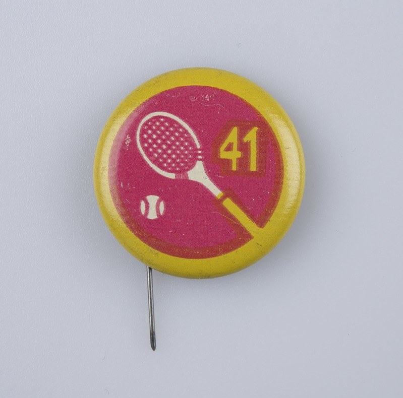 Gelb-rosa Abzeichen mit Tennis-Schläger-Design, befestigt an einer weißen Oberfläche mit einer Nadel, mit Text und Zahlen.