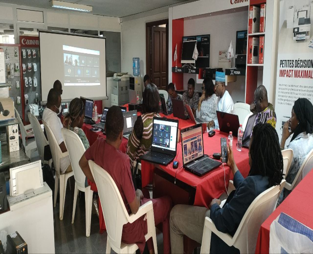 Gruppe von Menschen an einem Tisch mit Laptops und anderen Gegenst├Ąnden bei einem digitalen Marketing-Workshop, mit einem Banner, einem Display und Deckenleuchten im Raum.
