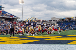 Ein Fußballspiel im Gange mit Spielern in Helmen im Vordergrund, Zuschauern und Stadionelementen im Hintergrund, mit Text unten im Bild.