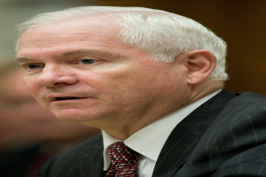 Ein Mann in einem schwarzen Blazer, weißem Hemd und roter Krawatte sitzt an einem Tisch und sieht ernst aus, wobei er wie ein Generalstaatsanwalt mit einem unscharfen Hintergrund wirkt.